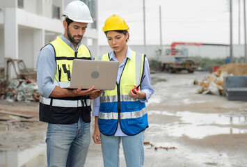Engineer man and female architect wear safety helmets discuss housing development project at construction site using laptop computer. Contractor manager examining building estate infrastructure.