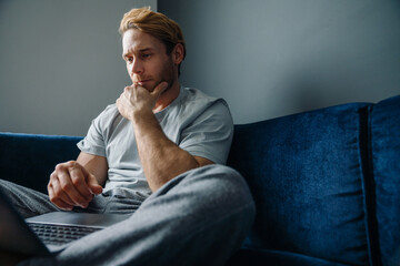 Young bearded man using laptop computer while sitting on sofa at home