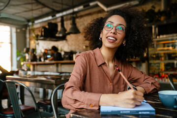 Young black woman making notes in planner book in cafe indoors