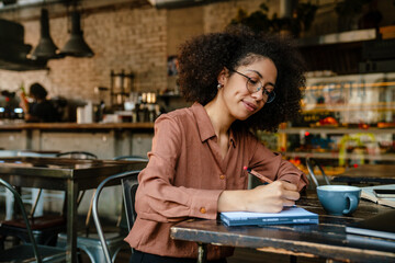 Young black woman making notes in planner book in cafe indoors