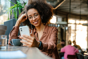 Young black woman using cellphone and earphones in cafe indoors