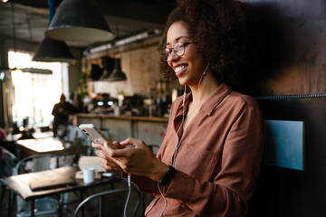 Young black woman using cellphone and earphones in cafe indoors