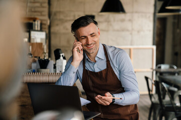 Adult white waiter using cellphone and laptop while working in cafe