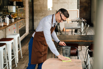 Adult waiter wearing apron and eyeglasses cleaning tables in cafe