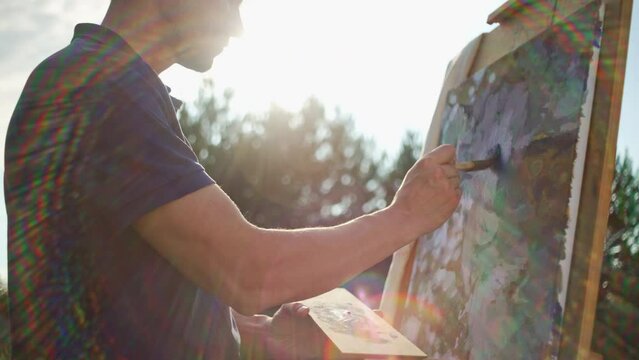 A male artist paints a picture on an easel with a brush. Beautiful sunset in nature, inspiration tree art and hobby drawing. High quality 4k footage