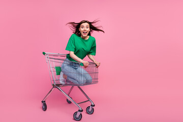Full length portrait of cheerful overjoyed girl sitting inside supermarket trolley isolated on pink...