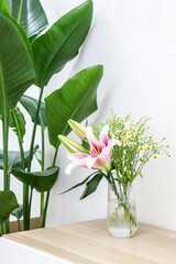 Vertical shot of bouquet of beautiful flowers (pink lilies and cute daises) in glass vase on wooden surface next to a Giant White Bird of Paradise (Strelitzia nicolai), decorating home interior