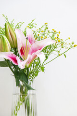 Vertical shot close up of bouquet of flowers in glass vase, showing a blossoming pink lily, two unopened bulb-shaped lilies, and small daises, on left side of frame against white background