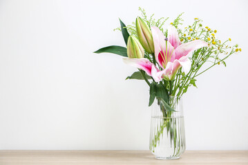 Bouquet of beautiful flowers in glass vase, blossoming pink lily, two opened bulb shaped lilies, and small daisies - decorating right side of wooden desk against white background
