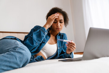 Young woman holding pregnancy test and using laptop in bed at home