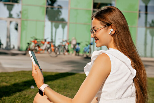 Profile close up portrait of smiling glad woman with loose hair wearing summer dress sitting on the lawn in morning in headphones. Woman with smartphone in headphones on quay 