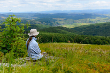 A man with long hair and a hat is sitting on a rock and admiring the mountain landscape