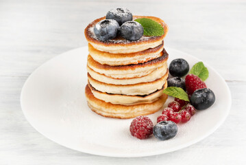 Pancakes with powdered sugar, blueberries, currants and raspberries on a white plate on a light table.
