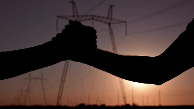 Handshake of two electrical engineers against the backdrop of high-voltage electrical tower at sunset. Silhouette hands close up. Teamwork, signing contract, agreement between businessmen Slow motion
