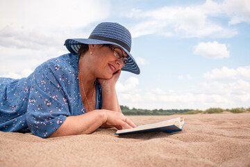 Elderly woman reads a book on the sandy shore.