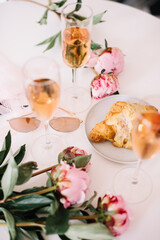 Delicious rose wine in glasses, pink peonies and a croissant on the white table background, flat lay, top view