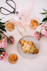 Delicious rose wine in glasses, pink peonies and a croissant on the white table background, flat lay, top view