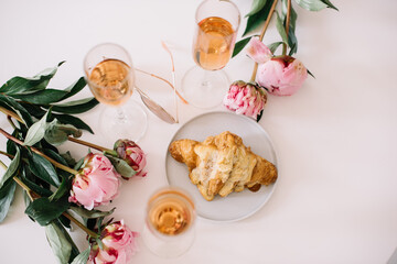 Delicious rose wine in glasses, pink peonies and a croissant on the white table background, flat...