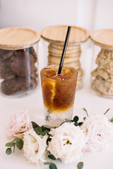 Delicious refreshing ice cold espresso tonic cocktail standing on the white table with three cookie jars on the background and blossoming flowers on the foreground, close up vertical photo