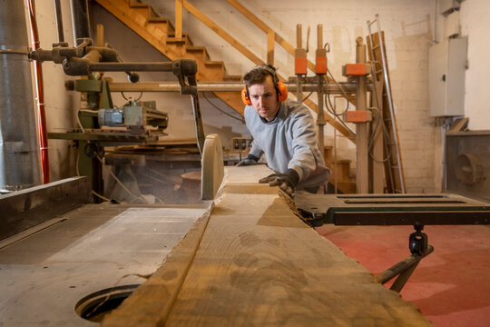 Young carpenter boy, concentrated on his job while cutting a long plank of wood with a circular saw.