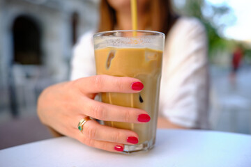 Iced coffee with milk. Close-up of a woman's hand holding a coffee with a straw and ice. Hand with red nails. Iced coffee being drunk in a cafe outside on a summer sunny day. 