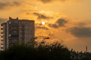 Orange sky at sunset, a high-rise building and the sun nearby, Clouds and trees.