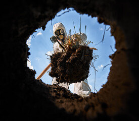 View from inside pit of ecologist digging pit by shovel and planting tree, wearing gas mask and...