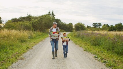 Fototapeta premium Farmer and his son in front of a sunset agricultural landscape. Man and a boy in a countryside field. Fatherhood, country life, farming and country lifestyle.