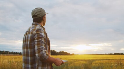 Farmer with a tablet computer in front of a sunset agricultural landscape. Man in a countryside...