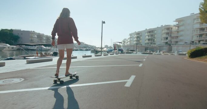 Young skinny woman cruises on longboard in front of yachts and houses in early summer morning. Woman practice skateboarding. Handheld footage with lens flare in frame. Summertime and youth freedom