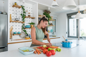 Young sporty woman cutting and cooking fresh natural vegetables at kitchen and cooking for healthy lunch. Self care yoga nutritionist girl having healthy meal after workout.