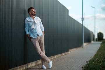 Fashion model handsome young man with trendy hairstyle in sunglasses in white shirt in light jeans posing near vintage wall on street on sunny day. Attractive american guy on walk. Copy space.