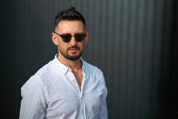 Obraz premium Fashion model handsome young man with trendy hairstyle in sunglasses in white shirt in light jeans posing near vintage wall on street on sunny day. Attractive american guy on walk. Copy space.