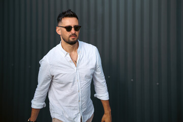 Obraz premium Fashion model handsome young man with trendy hairstyle in sunglasses in white shirt in light jeans posing near vintage wall on street on sunny day. Attractive american guy on walk. Copy space.
