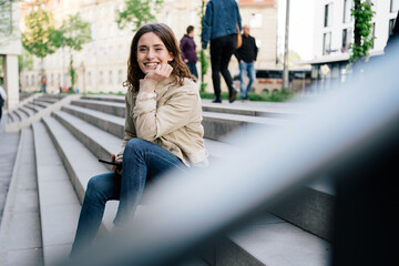 Young woman with cell phone sits on stairs and looks at camera laughing