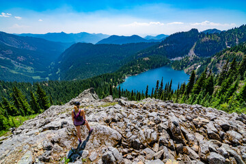 Fototapeta na wymiar Adventurous athletic woman standing on a mountain top overlooking an alpine lake and a rugged mountain range on a beautiful sunny day in the Pacific Northwest.