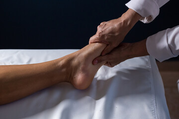 Detalhe das mãos de massagista aplicando massagem terapêutica no pé de um paciente que está deitado em uma maca com lençol branco. - obrazy, fototapety, plakaty