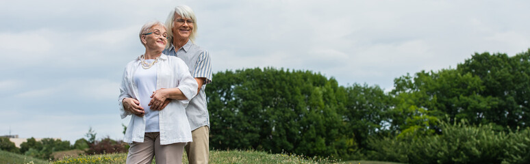 Obraz premium cheerful senior man in glasses hugging wife with grey hair and standing outside, banner.