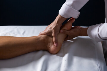 Detalhe das mãos de massagista aplicando massagem terapêutica no pé de um paciente que está deitado em uma maca com lençol branco. - obrazy, fototapety, plakaty