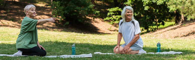 full length of senior woman pointing with finger near happy husband on fitness mat in green park, banner.