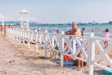 Old white woman, in a bathing suit and a bandana stands leaning on a fence behind the Black Sea and...