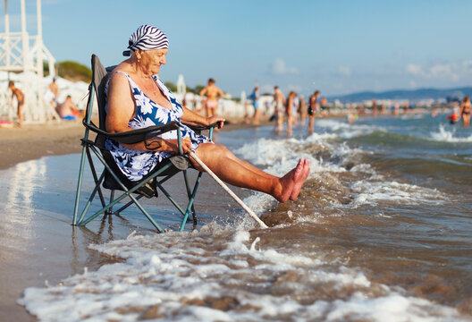 Old white woman in a swimsuit and a bandana sits in a tourist chair near the Black Sea near the city of Anaps and meets the oncoming wave