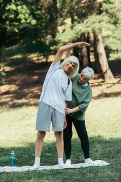 senior woman training happy husband with grey hair standing on fitness mat in park.