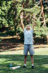 full length of happy senior man in sportswear exercising near sports bottle and fitness mat on grass.