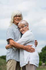 happy senior husband and wife with grey hair embracing against cloudy sky.