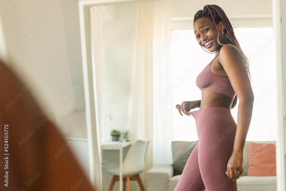 Wall mural Fit black lady in front of mirror checking training and dieting results, looking at herself and smiling