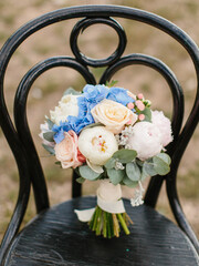 A delicate bridal bouquet of white peonies, cream roses, blue hydrangea, and eucalyptus leaves, tied with a white ribbon, stands on a black wooden chair.