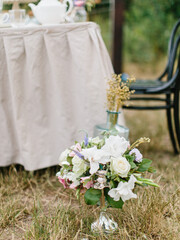 A bouquet of white and lilac eustoma, pink roses, twigs, and leaves stand in a copper candlestick on the grass. In the background is a banquet table with a white tablecloth.