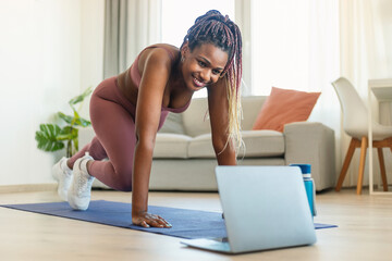 Young african american fitness lady doing leg exercises and watching online workout on laptop at home
