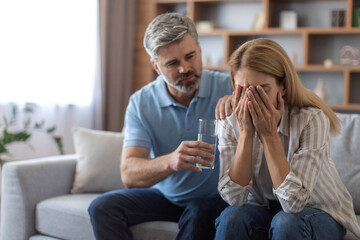Serious mature caucasian husband calm upset crying wife, gives glass of water, lady suffers from pain and depression
